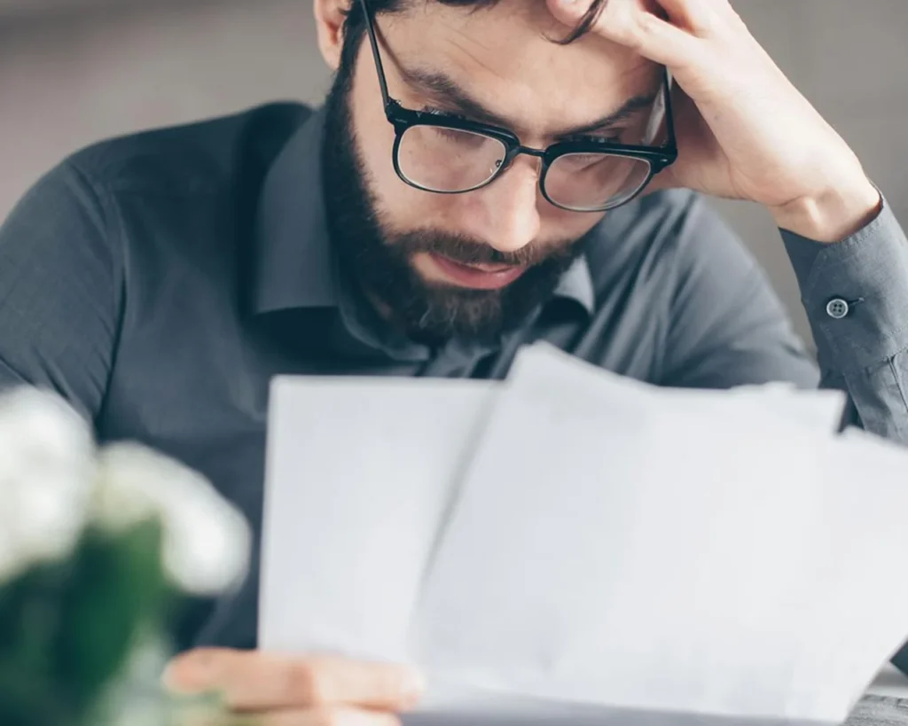A man with glasses and a beard is intently reviewing paperwork at a desk.