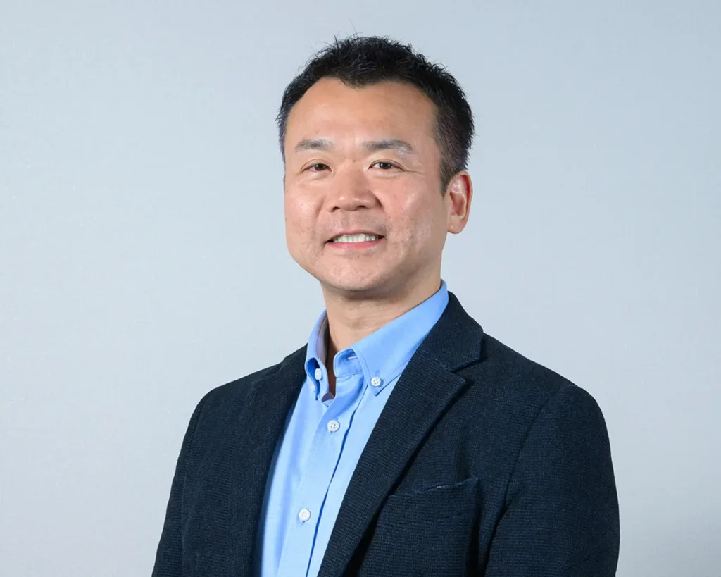 A man in a blue shirt and jacket stands against a gray wall, looking confidently at the camera.